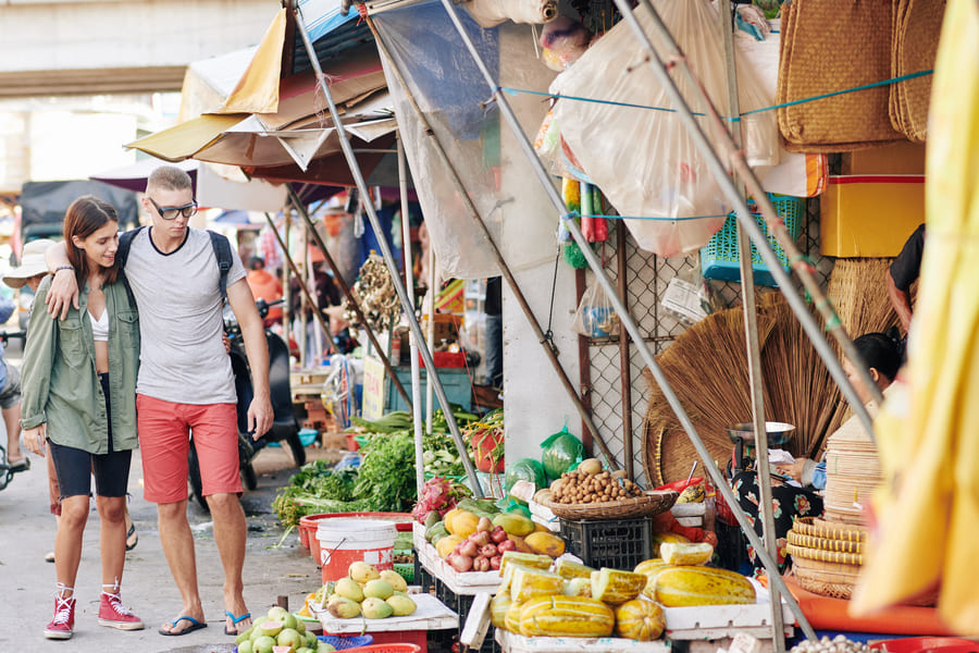 Pareja feliz caminando juntos en un mercado al aire libre en China, con puestos llenos de frutas y verduras a su alrededor.