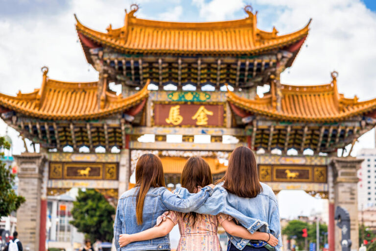 Tres jóvenes posan alegremente frente a una puerta china, rodeadas de un ambiente cultural vibrante.