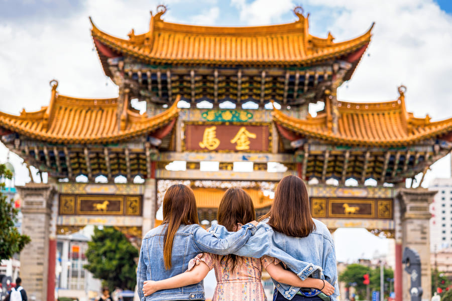 Tres jóvenes posan alegremente frente a una puerta china, rodeadas de un ambiente cultural vibrante.